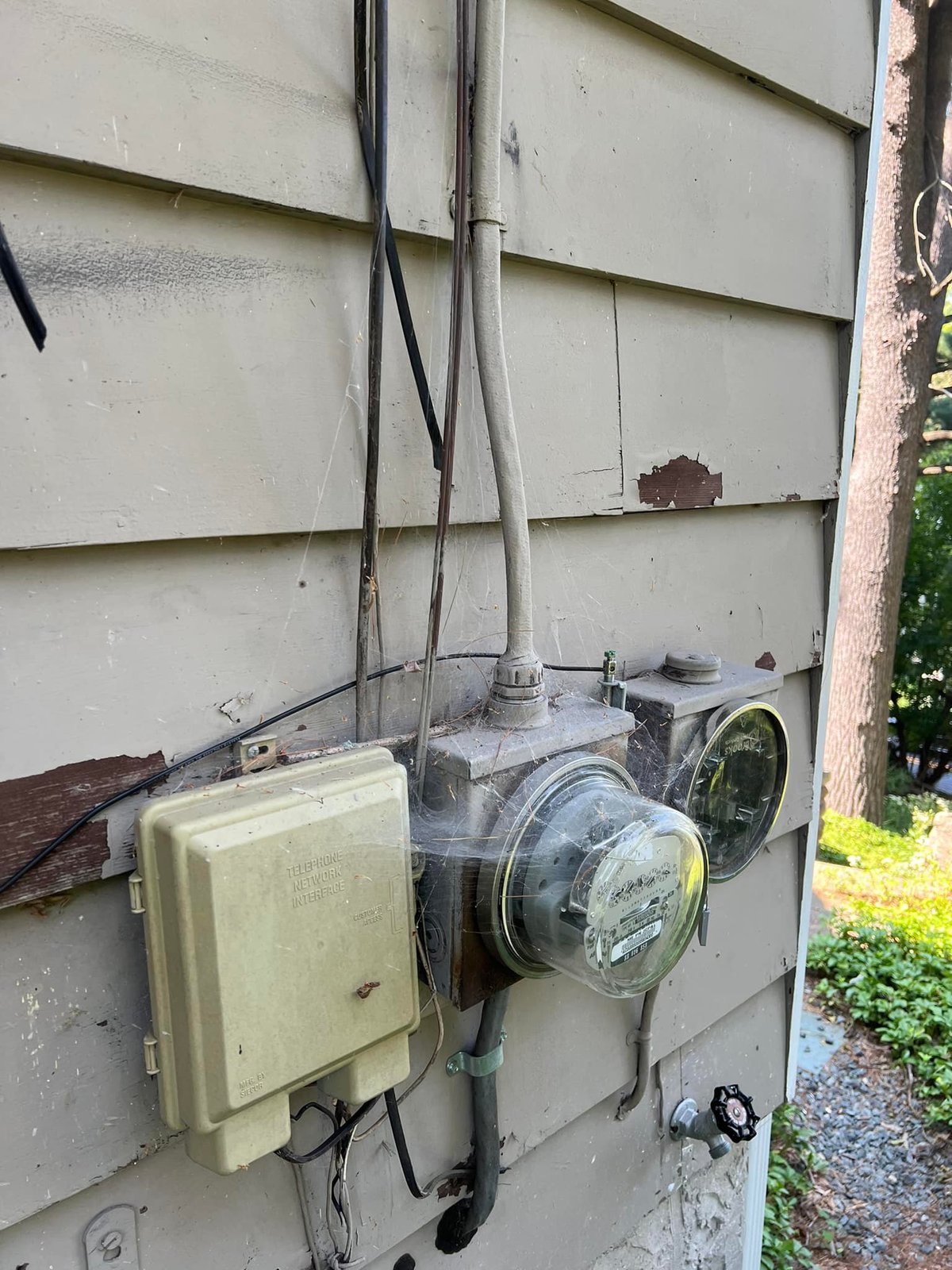 Old electric meter and the network interface on weathered siding. Cobwebs, peeling paint, and the bottom of the meter base rotted out.